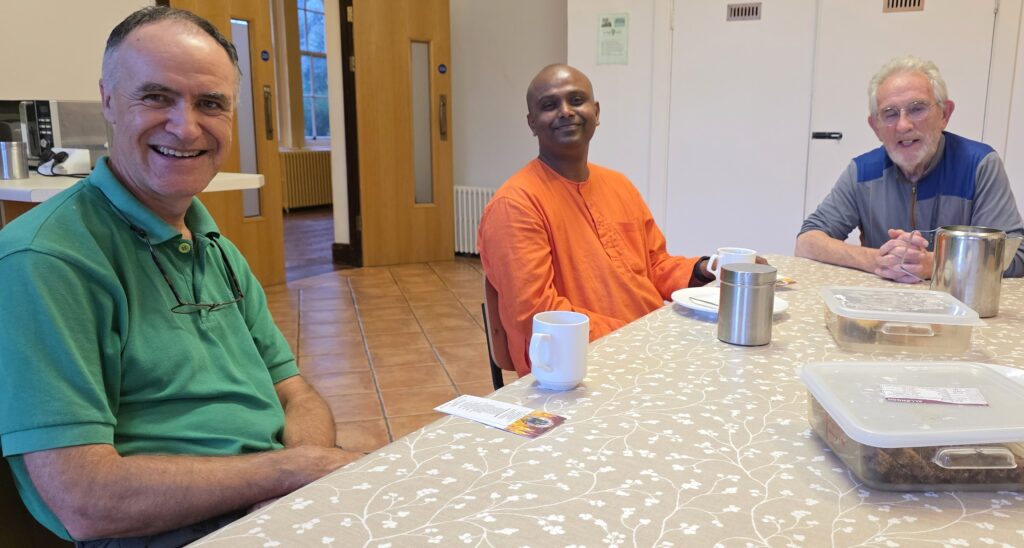 Three men sit at a sunlit table in a community room, smiling at the camera with mugs and a plastic lunch box on the table.