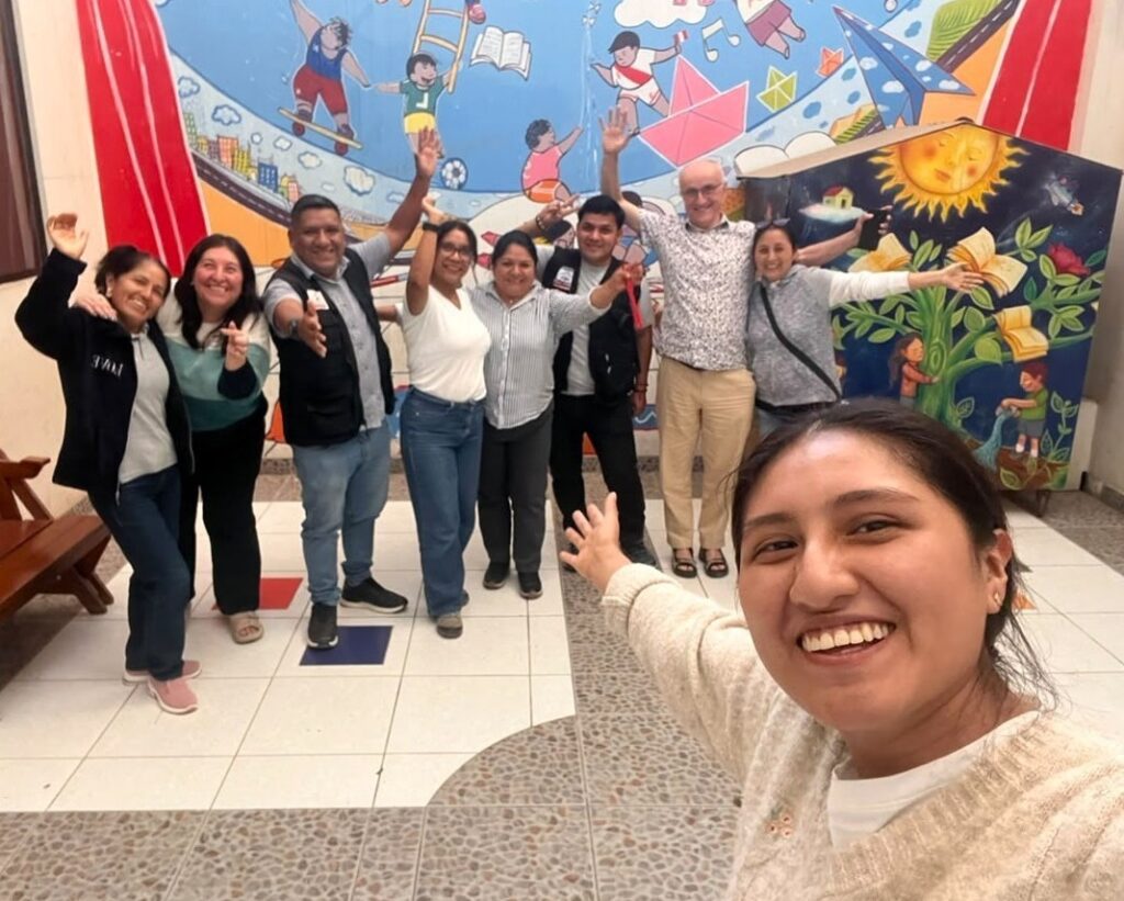 Group of diverse adults posing together in a hallway, arms raised in celebration, with a colorful mural behind them; a woman in the foreground takes a selfie.