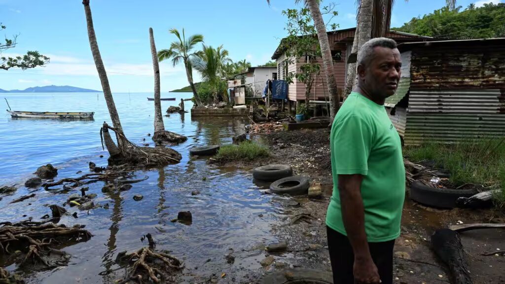 Man in a bright green shirt stands by a muddy shoreline with palm trees, weathered houses, and discarded tires near the water.