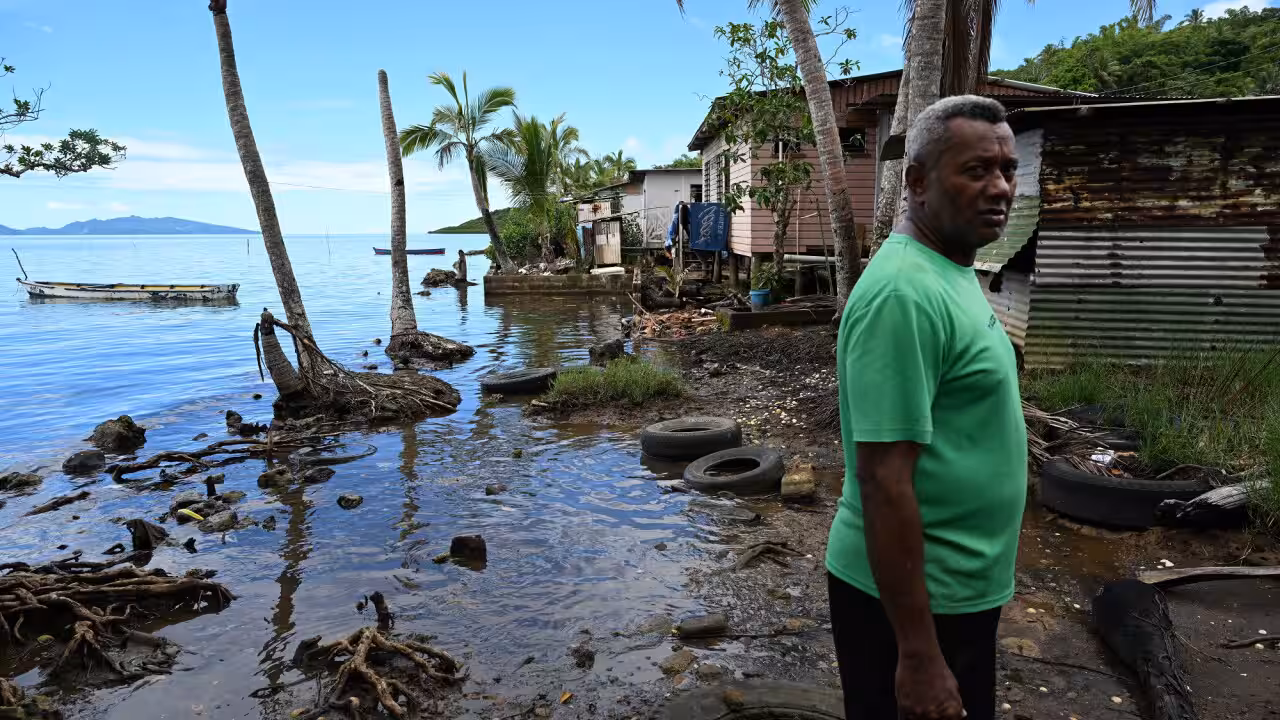 Man in a bright green shirt stands by a muddy shoreline with palm trees, weathered houses, and discarded tires near the water.