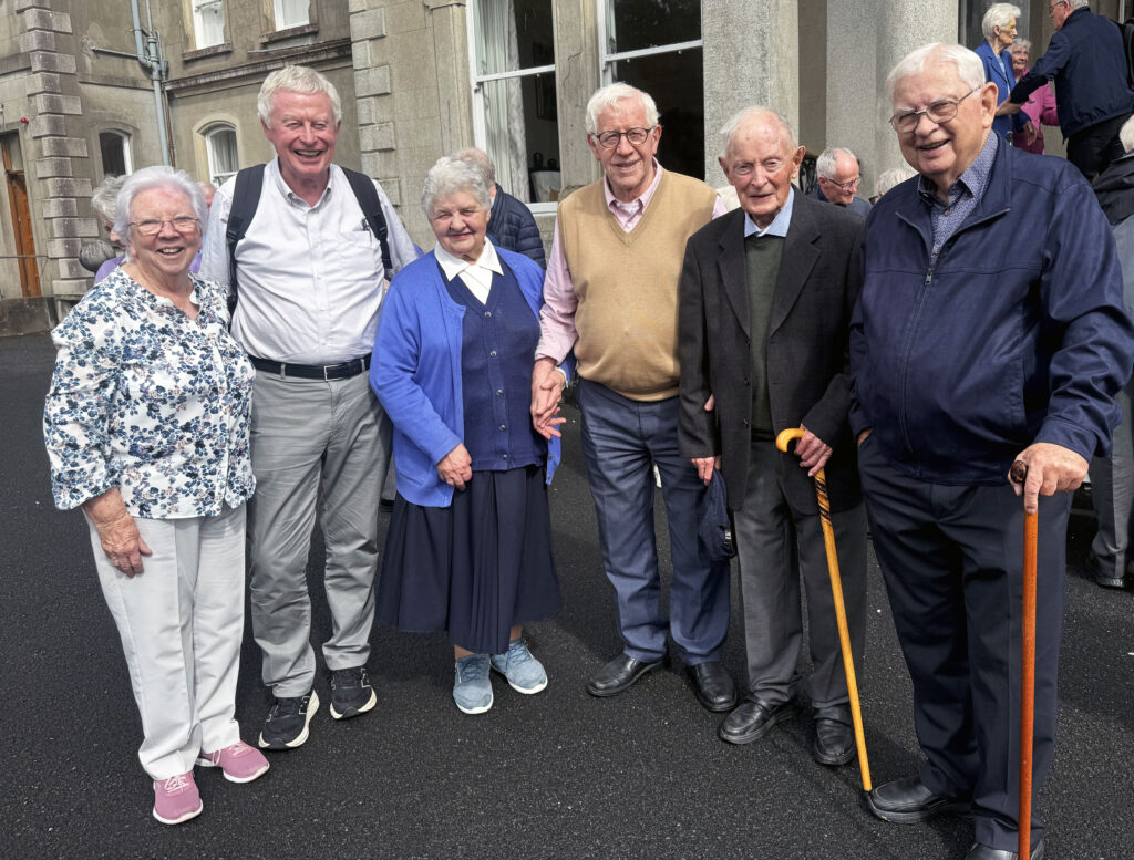 Group of smiling elderly people standing together outside a stone building on a paved street, some with canes and wearing smart casual clothes.