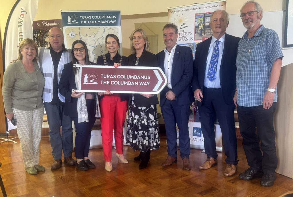 Eight people stand indoors, smiling, holding a burgundy sign that reads 'TURAS COLUMBANUS THE COLUMBAN WAY' with banners and a map behind them on display boards in a conference setting.