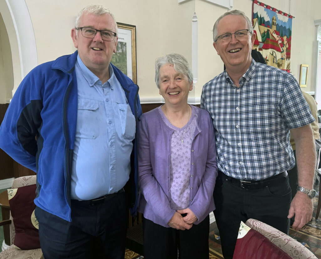 Three smiling adults pose together indoors, center woman with short gray hair wearing a lavender cardigan flanked by two men in blue and checkered shirts.