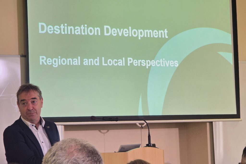 Speaker in a dark suit presenting at a conference, with a green slide reading 'Destination Development' and 'Regional and Local Perspectives' behind him.