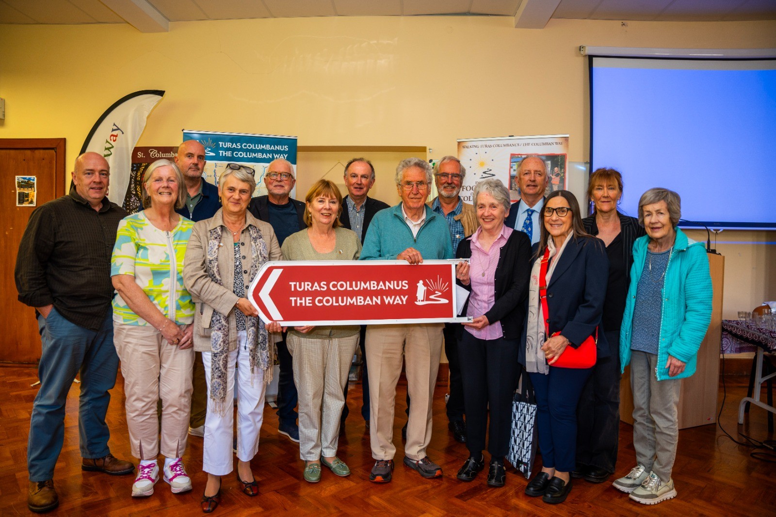 Group of diverse adults indoors posing for a photo, holding a red sign that reads 'TURAS COLUMBANUS THE COLUMBAN WAY' at a community event with banners in the background.