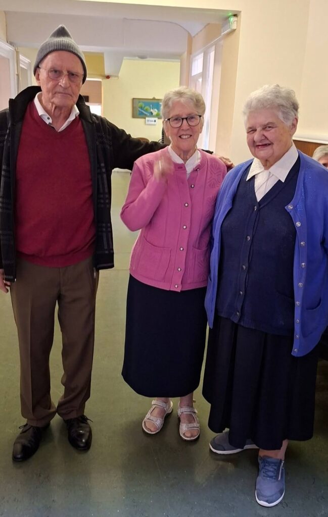 Three elderly friends posing together indoors, smiling at camera in a hallway.