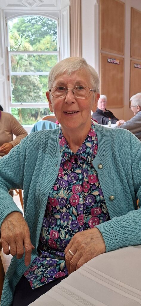 Older woman with short white hair and glasses sits at a table, wearing a turquoise cardigan and floral blouse in a bright dining room.