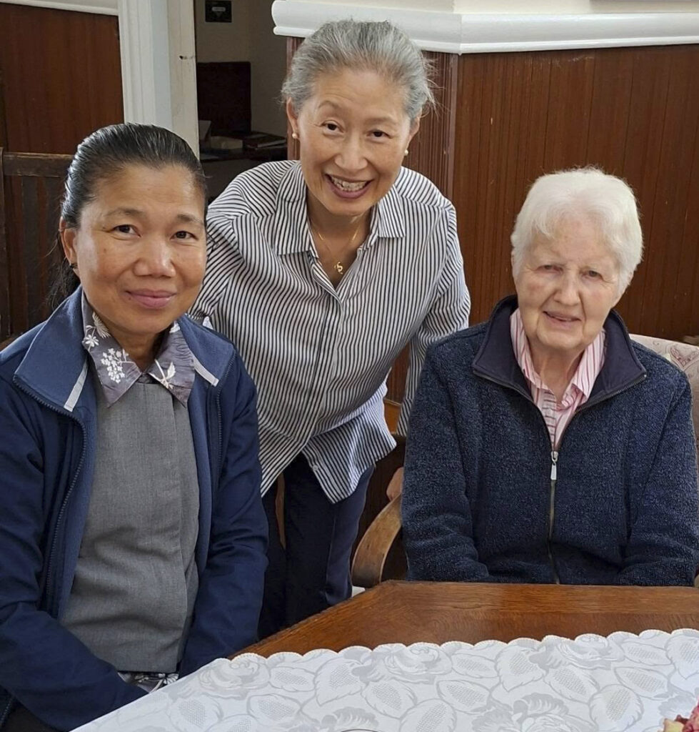 Three women of different ages sit together at a wooden table, smiling at the camera indoors.