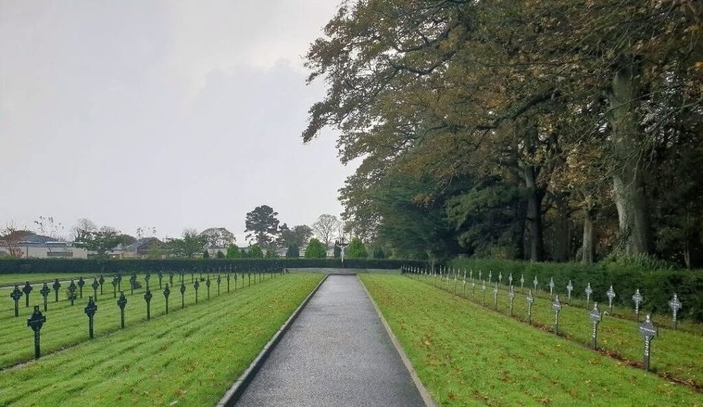 Grassy war cemetery with two rows of identical grave markers flanking a central asphalt path and a statue at the far end.