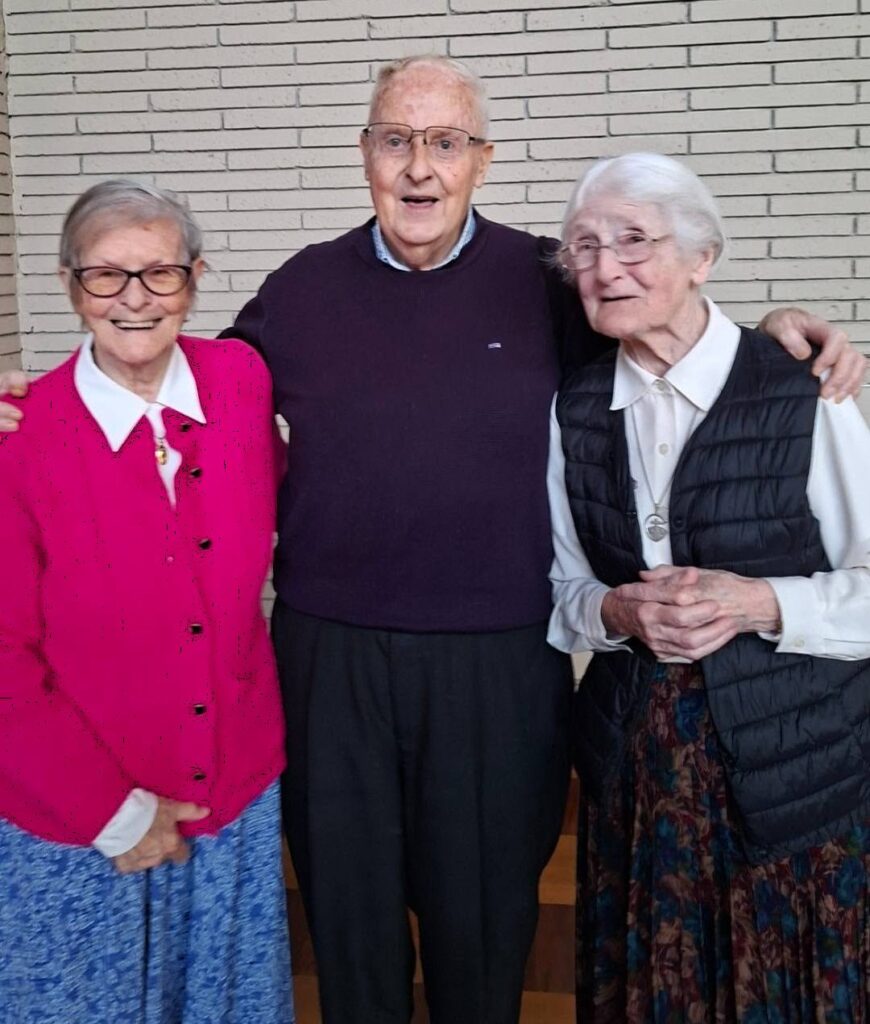 Three elderly adults standing together and smiling in front of a brick wall; left woman in a bright pink cardigan, center man in a purple sweater, right woman in a white blouse and black vest.