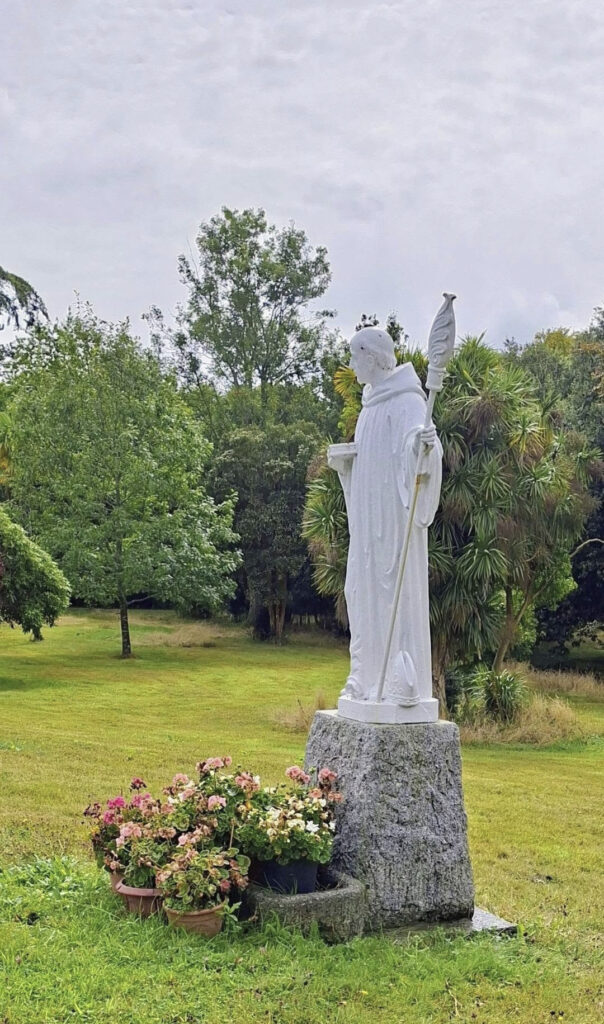 White statue of a robed saint holding a staff, standing on a stone pedestal in a garden with pink flowers nearby.