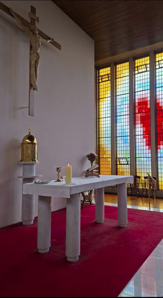 Altar in a church with a crucifix on the wall, a white marble table set with a candle, chalice, and open Bible, and colorful stained-glass windows in the background.