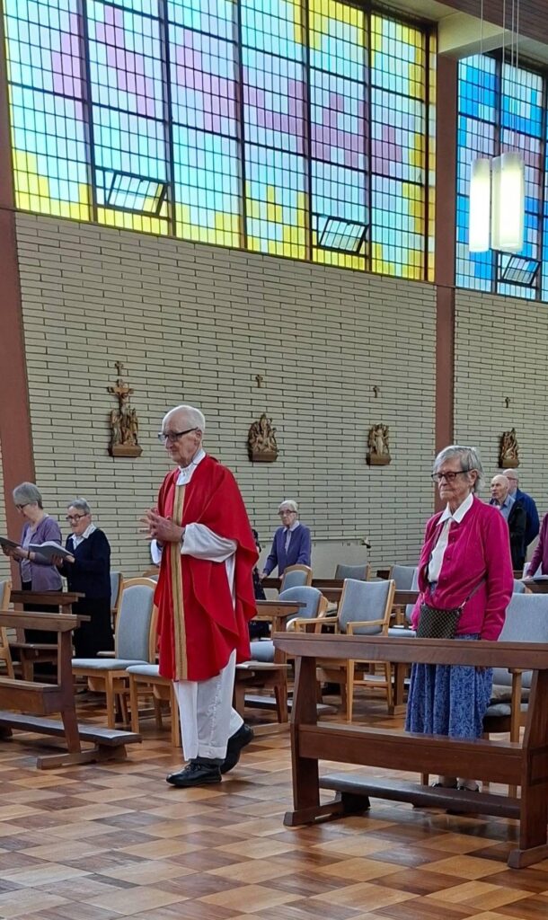 Priest in red liturgical vestments leads a service as parishioners stand in a chapel with wooden pews and vibrant stained-glass windows above.