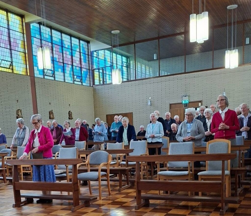 Group of elderly people standing in a church sanctuary during a service; colorful stained-glass windows and wooden pews surrounding them.