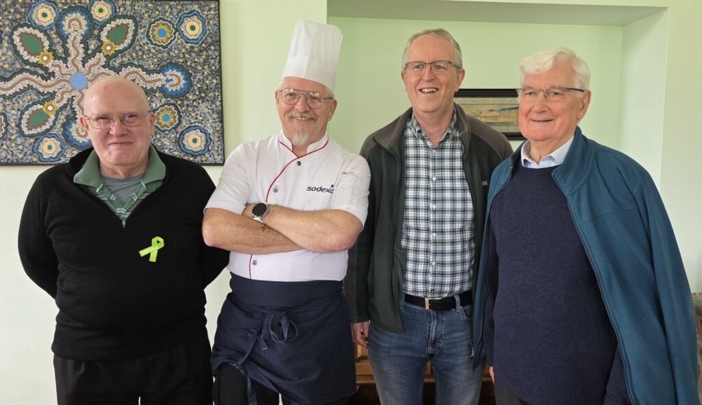 Four older men posing indoors; a chef in white uniform and hat stands second from left, others in casual clothes, against a green wall with art behind.