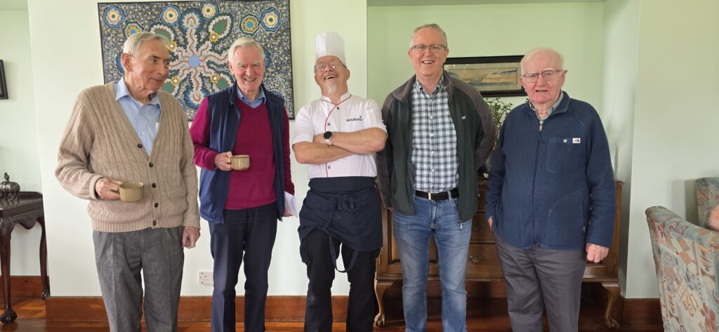 Five older men stand together in a living room, smiling for the camera; one wears a chef's hat and white uniform in the center-right of the group.