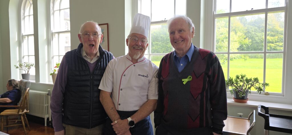 Three elderly men posing and smiling indoors near a large window, with the center man wearing a chef's hat and white uniform.