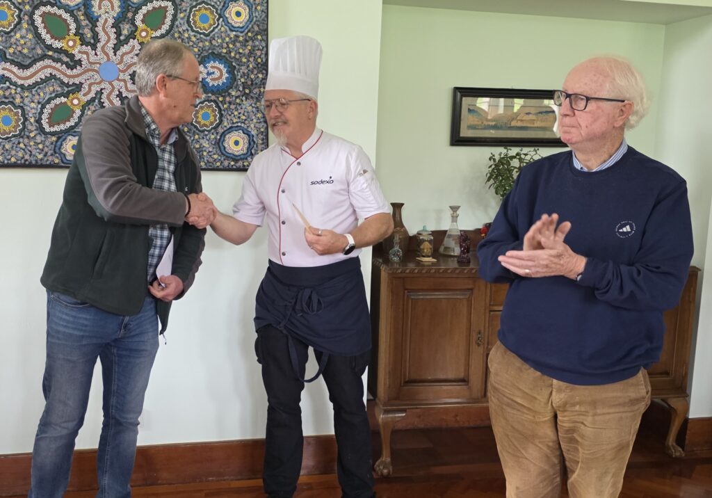Three men indoors: two shaking hands while a third man claps nearby; a chef stands between them in a room with a decorative wall hanging.
