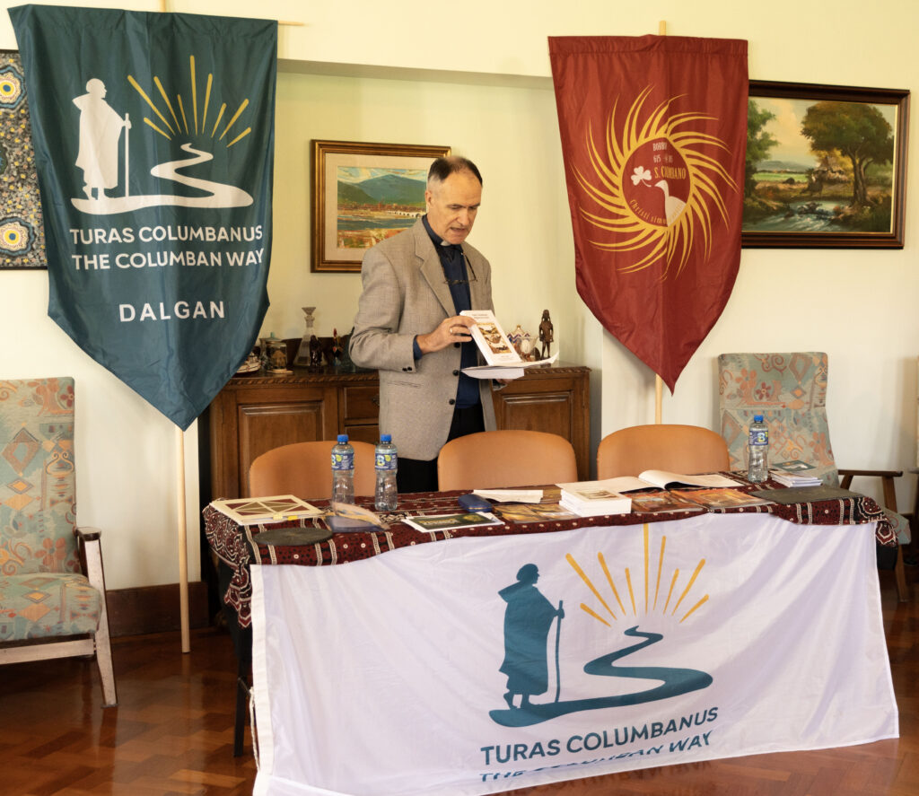 Man in a gray blazer reads from papers at a table, flanked by a blue Turas Columbanus banner and a red banner with a sun emblem.