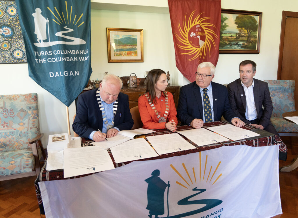Four people in business attire sign documents at a ceremonial signing table, with teal and red banners behind them.