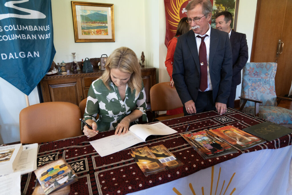 Woman signs documents at a table while a man in a suit stands beside her in a formal office setting with banners and art in the background.