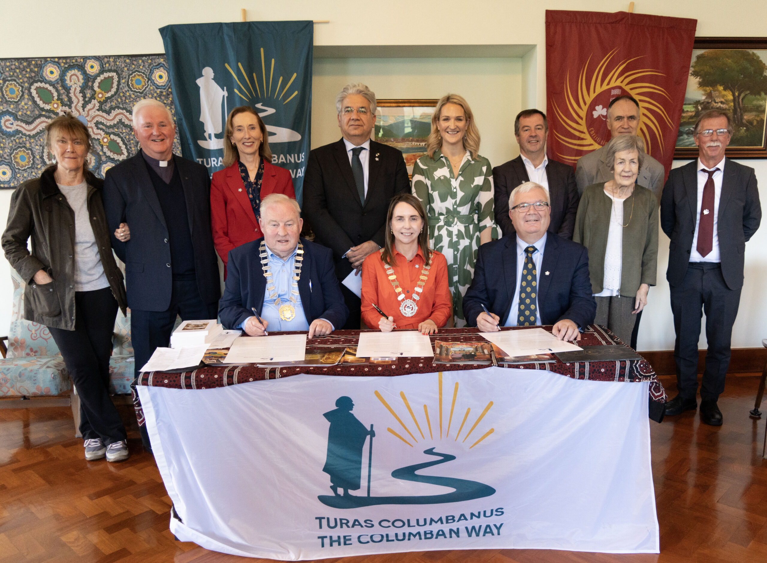 Group of ten adults at a signing ceremony, two seated signing documents at a table, banners behind them.
