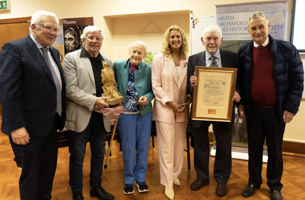 Six adults pose indoors with awards: one man holds a carved statue, another a framed certificate, with a Meath Archaeology banner in the background.