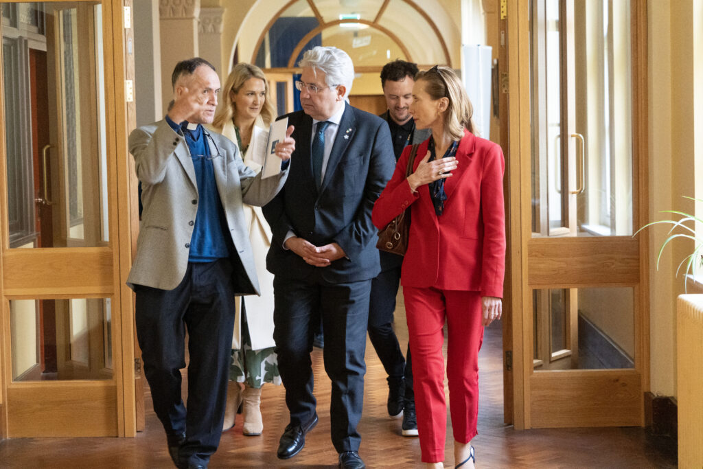 Group of five adults walking down a wooden hallway; front man in a gray blazer and clerical collar gestures, woman in light coat holds papers, others in business attire follow.