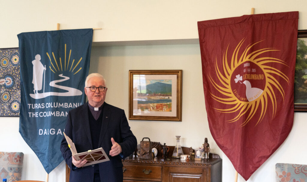 Older man in a dark suit and clerical collar speaks, holding papers, flanked by blue and burgundy banners in a room with a framed painting behind him.