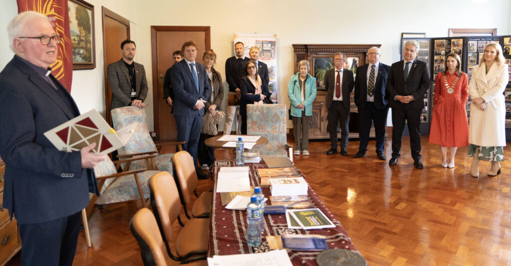 A group of formally dressed adults stands in a conference room beside a long table covered with pamphlets and bottles, listening to a speaker at the left foreground.