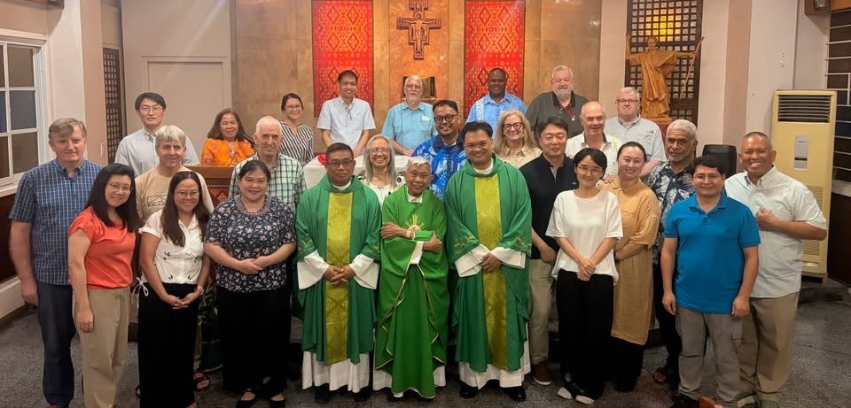 Group photo of about 25 people posing in a church hall, with three priests in green vestments at the front center and an altar backdrop behind them.